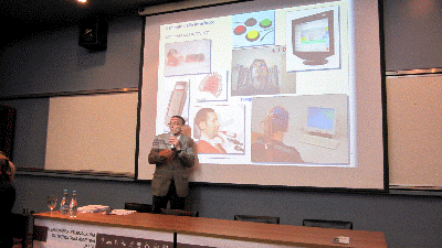Professor Renzo está em pé e fala ao microfone.  Ao fundo visualiza-se uma tela com imagens de recursos de tecnologia assistiva para acesso ao computador .